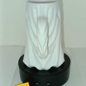 K&ouml;nnte beinhalten: Ein wei&szlig;er Keramikbecher in Form eines drapierten Geistes mit vertikalem Henkel. Der Becher steht auf einem schwarzen, rotierenden Pr&auml;sentationssockel. Ein gelber Zettel zeigt den Text 330 ml und 11,2 oz als Fassungsverm&ouml;gen.