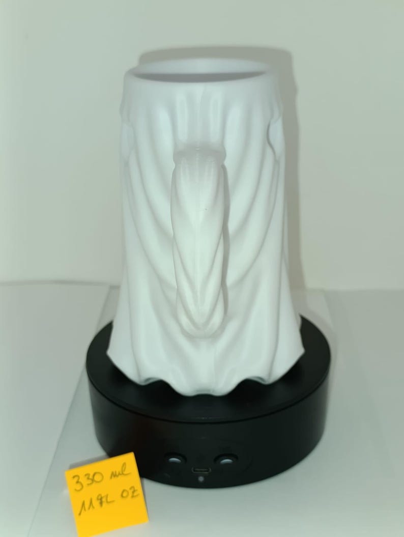 K&ouml;nnte beinhalten: Ein wei&szlig;er Keramikbecher in Form eines drapierten Geistes mit vertikalem Henkel. Der Becher steht auf einem schwarzen, rotierenden Pr&auml;sentationssockel. Ein gelber Zettel zeigt den Text 330 ml und 11,2 oz als Fassungsverm&ouml;gen.