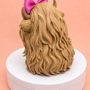 May include: A figurine of a Highland cow with long, textured light brown hair, two curved horns, and a bright pink satin bow attached between the horns. The figure is shown from the back, displaying the detailed, flowing strands of hair.