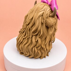 May include: A sculpted figurine of a Highland cow head with long, textured light brown fur, two curved dark brown horns, and a bright pink bow attached to the top. The sculpture is rendered in a realistic style with detailed hair strands.