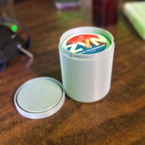 May include: A white cylindrical container with a lid, open to reveal a circular product with the text "COOL WATERMELON SLIM". The container is on a wooden surface.