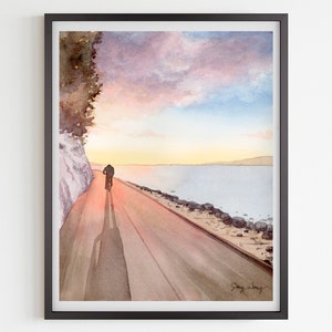 May include: Watercolour painting of a person cycling on a path beside a body of water at sunset. The sky is a mix of pink, purple and blue with a few clouds. The water is a light blue and the path is a light brown. The person's shadow is cast on the path.