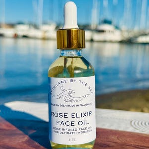 Op de afbeelding: Een glazen flesje met rozen-elixir gezichtsolie met een gouden dop en een wit etiket. Het etiket vermeldt "Skincare by the Sea" en "Rose Elixir Face Oil". De fles staat op een houten oppervlak.