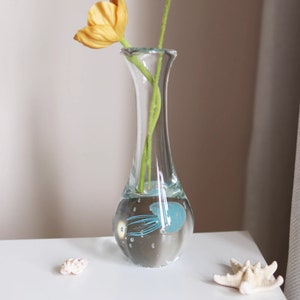 Könnte beinhalten: Eine klare Glasvase mit einem blauen Quallen-Design, gefüllt mit Wasser und zwei gelben Blumen.