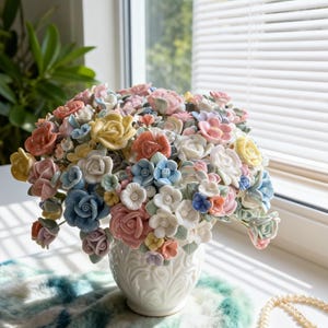 Puede incluir: Un arreglo floral de cerámica en un jarrón blanco. El ramo presenta una variedad de rosas de colores pastel y otras flores, incluyendo flores rosas, amarillas, azules y blancas. El jarrón tiene un diseño detallado y texturizado.