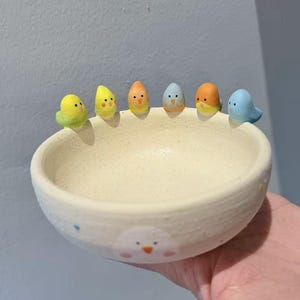 Puede incluir: Un cuenco de cerámica hecho a mano con un interior color crema y una fila de seis figuras de pájaros coloridos alrededor del borde. Los pájaros son amarillos, naranjas y azules. El cuenco tiene un lindo diseño de cara de pájaro en el exterior.