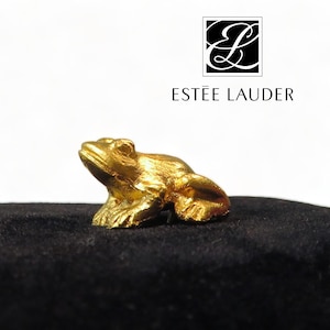 Peut inclure: Une petite figurine de grenouille dorée. La grenouille est en position assise, les pattes avant légèrement relevées. Le logo Estée Lauder est dans le coin supérieur droit.