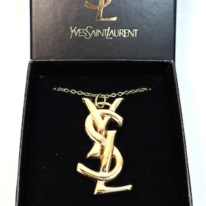 May include: A gold-toned metal YSL logo pendant necklace resting inside a black presentation box. The box lid features the gold YSL monogram and the text Yves Saint Laurent printed in gold. The pendant is a large, polished gold-tone metal logo.