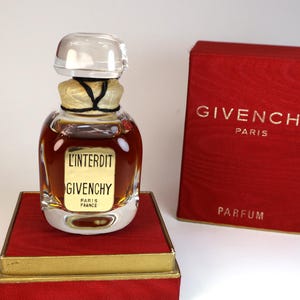 May include: A glass perfume bottle with a clear stopper and a gold label reading L'INTERDIT GIVENCHY PARIS FRANCE, filled with amber liquid. It sits on a red and gold box next to a matching red box with the text GIVENCHY PARIS PARFUM printed in gold.