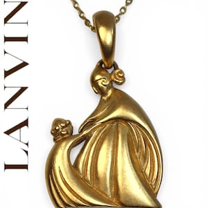 Puede incluir: Un collar con colgante dorado que presenta un diseño estilizado de madre e hijo. El colgante tiene un acabado mate y cuelga de una delicada cadena. La palabra "LANVIN" está impresa verticalmente en el lado izquierdo.