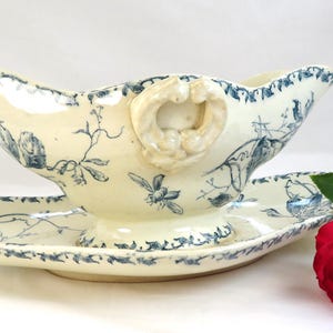 Puede incluir: Salsera y platillo a juego de color crema con estampado floral y de pájaros azul. La salsera tiene un asa decorativa y un borde festoneado. Una rosa roja con una hoja verde está en primer plano.