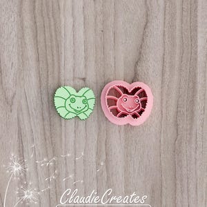May include: Two plastic cookie cutters, one green and one pink, shaped like a lizard with a smiling face.