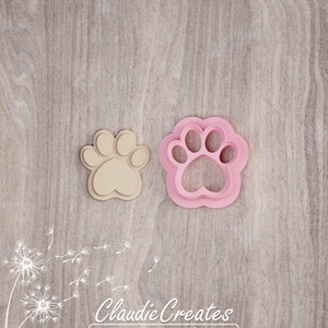 May include: A set of two cookie cutters, one pink and one beige, shaped like a paw print. The cutters are on a woodgrain background.