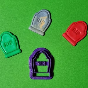 May include: Four tombstone-shaped cookie cutters, one purple, one green, one red, and one grey. The cutters are on a green surface. The purple cutter is the largest and has the word "RIP" inscribed on it. The other cutters have the word "RIP" inscribed on them as well.