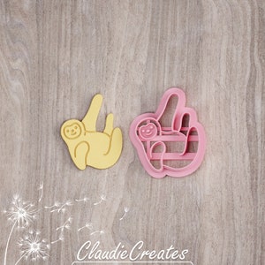 May include: Two cookie cutters, one yellow and one pink, shaped like a sloth. The yellow cutter is a simple outline of a sloth, while the pink cutter has a striped pattern.