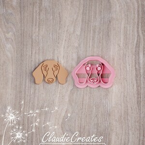 May include: Two dachshund dog cookie cutters, one brown and one pink. The cutters are on a woodgrain background.