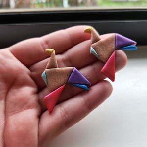 May include: Two origami-style bird earrings made of colourful polymer clay. The birds are folded into a geometric shape and have a glittery finish. The earrings are a light brown colour with accents of purple, blue, red, and gold.