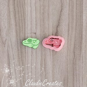 May include: Two plastic cookie cutters shaped like lizard heads. The cutter on the left is green and the cutter on the right is pink.