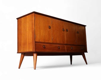 May include: A mid-century wooden cabinet in a warm brown finish. It has four doors, two drawers, and rounded wooden pulls. The cabinet is supported by four angled legs. The cabinet's dimensions are approximately 150 cm in width.