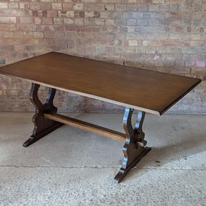 May include: A rectangular wooden dining table with a smooth, dark brown finish. The table features a trestle base design with two curved, vertical supports connected by a horizontal stretcher bar that runs between the two legs.