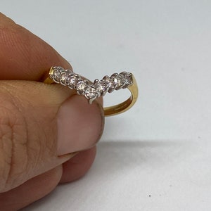 May include: A gold-toned ring with a V-shaped design, set with multiple clear gemstones. The ring is held against a white background, showcasing the sparkle of the stones. The band appears to be a classic style, suitable for various occasions.