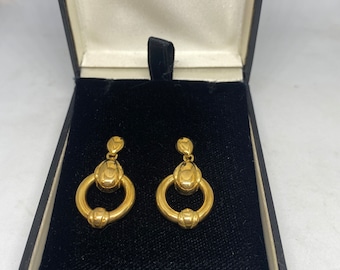 May include: A pair of gold-colored earrings displayed in a black jewelry box. Each earring features a small top element, a rounded central design, and a circular bottom. The earrings are presented against a black velvet interior.