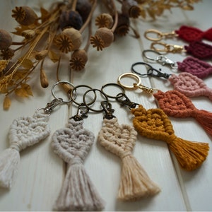 Op de afbeelding: Een set van acht handgemaakte macramé sleutelhangers met kwastjes in verschillende kleuren, waaronder wit, beige, oranje, rood en roze. Elke sleutelhanger heeft een hartvormig ontwerp en is bevestigd aan een zilveren sleutelring.