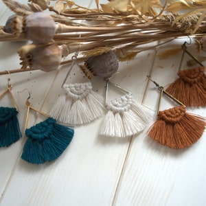 May include: Three pairs of triangle-shaped earrings with macrame fringe. The earrings are in white, teal, and brown.