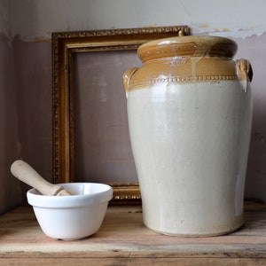 May include: A large, white ceramic jar with a brown lid and handles sits on a wooden shelf next to a white ceramic bowl and a wooden pestle. A gold picture frame is behind the jar.