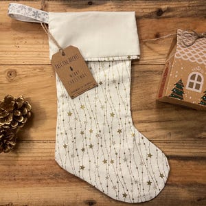 May include: White Christmas stocking with a gold star and string pattern. The stocking has a brown tag that reads "Past the Arches Merry Christmas Handmade in Croatia".