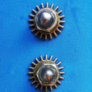 May include: Two silver metal gears with gold accents on a blue background. The gears have a starburst shape with a rounded centre.