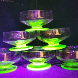 May include: Six clear glass dessert bowls with a green base. The bowls are stacked in a pyramid shape. The bowls are made of uranium glass and glow green under a black light.