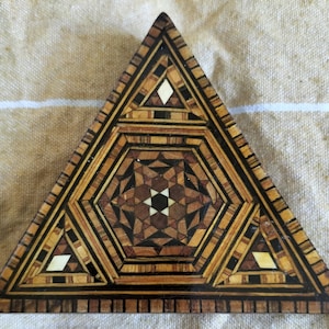 May include: A triangular wooden box with intricate geometric patterns inlaid in wood and ivory. The box is made of various shades of brown wood and has a geometric design.