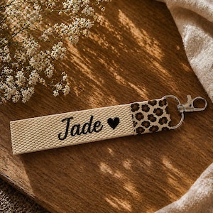 Peut inclure: Un porte-clés en tissu rectangulaire composé d'une sangle tissée couleur crème avec le prénom Jade et un cœur noir brodés en écriture cursive. L'extrémité présente un motif léopard marron et noir et un mousqueton en métal argenté.