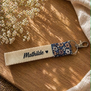 Peut inclure: Un porte-clés personnalisé avec le nom "Mathilde" en écriture cursive et un petit symbole de cœur. Le porte-clés est composé d'une sangle en tissu beige tissé avec un motif floral bleu, blanc et rose. Il possède un fermoir argenté.