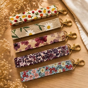 Peut inclure: Cinq porte-clés en tissu avec des fermoirs dorés. Les porte-clés présentent divers motifs floraux et fruités, notamment des marguerites, des fleurs violettes et un mélange de citrons, d'oranges et de fraises. Ils sont disposés sur une surface en bois.