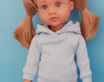 May include: A doll with blonde pigtails wearing a light blue hoodie and matching sweatpants. The doll has blue eyes and is standing against a solid blue background. The doll's outfit includes a front pocket on the hoodie.