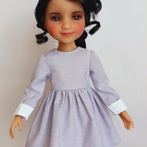 Puede incluir: Una muñeca con un vestido lavanda con puños blancos y botas plateadas. La muñeca tiene el pelo oscuro y los ojos marrones.