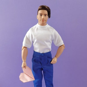 May include: A doll wearing a white t-shirt, blue trousers, and blue shoes. The doll is holding a pink origami heart.