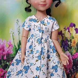 Peut inclure: Une poupée vêtue d'une robe blanche ornée d'un motif floral bleu. La robe a des manches courtes et une fermeture boutonnée. La poupée a les cheveux foncés et porte des chaussures grises. L'arrière-plan est une scène de jardin colorée.