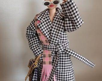 May include: A fashion doll wearing a black and white houndstooth coat with a pink scarf and a matching purse. The doll is also wearing white sunglasses and has blonde hair.