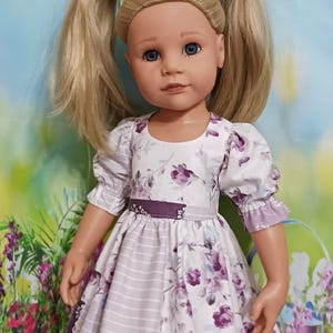 May include: A doll with blonde hair styled in pigtails, wearing a white dress with a purple floral pattern and a purple belt. The dress has short sleeves and a striped skirt. The doll is wearing white shoes.