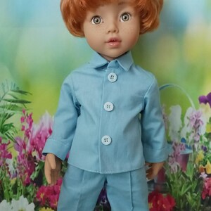 May include: A doll with red hair and freckles wearing a light blue pajama set. The outfit includes a button-down shirt and matching pants. The doll is wearing white shoes. The background features a floral design.