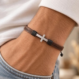 May include: A brown leather bracelet featuring a silver-toned metal cross charm at the center. The leather band is engraved with the text Matthew 6:34 on one side and Confirmed in Christ on the other, secured by black cord wrappings.