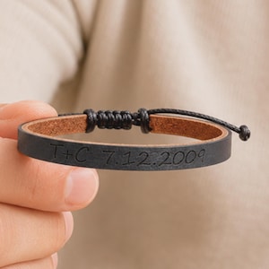 May include: A black leather bracelet featuring a tan interior and an adjustable black cord closure. The exterior surface of the leather band is engraved with the text T+C 7.12.2009 in a simple, dark font, creating a personalised accessory.