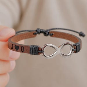 Può includere: Un bracciale in pelle marrone con un simbolo dell'infinito color argento al centro. Il cinturino in pelle è inciso con le lettere J e B separate da un'icona a forma di cuore. Il bracciale presenta dettagli in corda nera e una chiusura a nodo scorrevole.