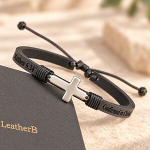 May include: A black leather bracelet featuring a silver-toned metal cross centerpiece. The leather band is engraved with the text Matthew 6:34 and Confirmed in Christ. The bracelet includes black cord accents and an adjustable sliding knot closure.