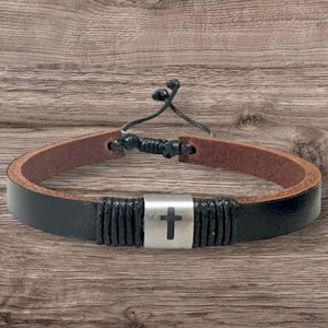 May include: Black leather bracelet with a brown interior. The bracelet features a silver-colored rectangular centerpiece with a cross symbol. The bracelet is adjustable with a black cord.