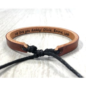May include: Brown leather bracelet with black cord closure. The bracelet is engraved with the text "We love you daddy! Olivia, Emma, Leah."