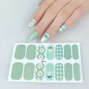 May include: A sheet of nail wraps with a variety of designs, including solid light green, daisy patterns, cherry blossom designs, and a gingham pattern. The nail wraps are shown with a hand that has matching nail art. The text "ZA513" is printed on the sheet.